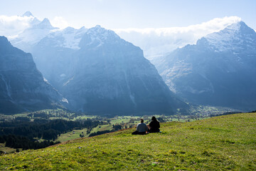 Fototapeta na wymiar A beautiful Alpine landscape with two tourists sitting on a green meadow hills admiring the distant hilly areas with pine trees below and fantastic mountain ranges covered with clouds on a clear day.