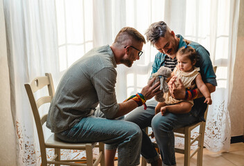 Male gay couple with adopted baby girl at home - Two handsome fathers playing with their daughter -...