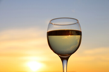 Glass with white wine on sunset background, evening sun and sky with clouds are reflected in the glass. Concept of celebration, wine industry