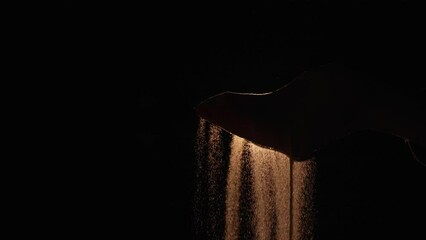 The dark silhouette of a female hand spilling sand from the palm of her hand on a black background with backlight. Particles of sand pour through fingers in the dark. Close up. Slow motion.