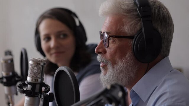 man and a woman record a podcast or interview with a microphone in a recording studio. an adult gray-haired man and a young European woman create audio content, record ads and audio books. adult