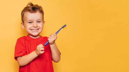 The happy kid is holding a blue toothbrush on a yellow background and smiling showing his teeth....