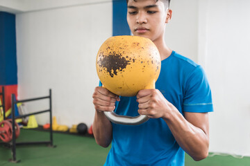 A young asian man grasps firmly an old rusted kettlebell. Holding upside-down. Preparing to do a set of goblet squats. Working our at the gym.