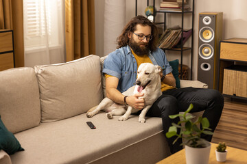 A bearded man spends his free time with his dog at home. The boy is holding a phone in his hands...