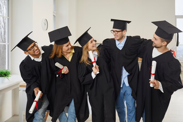 Group of happy university students celebrating graduation. Indoor shot of cheerful optimistic multiethnic classmates and friends in graduate academic hats and robes hugging and having fun in classroom