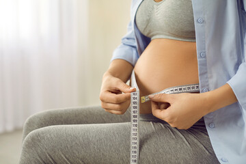 Pregnant woman measuring her big bare belly with a centimeter tape, close up. Happy mom-to-be in...
