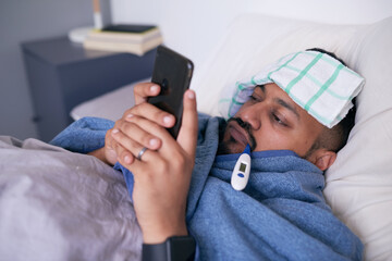 A sick man in bed with a thermometer looks at his mobile device
