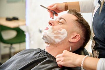 Young man shaving with straight edge razor by hairdresser in barbershop