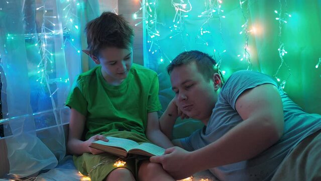 dad and son are reading a book lying on the couch. into the light of the garlands. bedroom decoration. a beautiful holiday