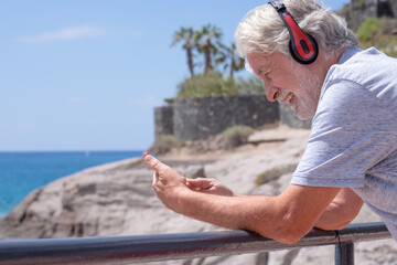 Smiling caucasian grandfather in outdoors at sea enjoying sunny day and vacation. Senior man with red headphones using modern technologies and wireless connection. Horizon over water and blue sky