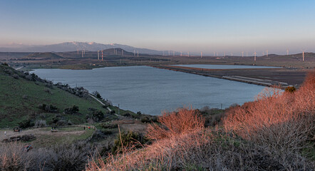 View of Bental water reservoir, the Golan hills with wind turbines and the Hermon Mounrain Range at sunset as seen from Mount Bental summit, Golan Heights, Israel.