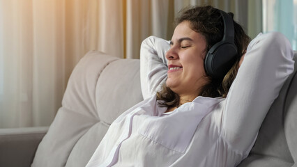 Young disabled long haired woman with cerebral palsy relaxes smiling and listening to music in headphones leaning back on sofa at home closeup. - obrazy, fototapety, plakaty