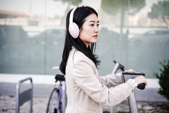 Chinese Business Woman Renting Electric Scooter In City,wearing Headphones.sustainable Transport
