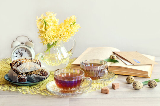 Spend time with friends: a bouquet of yellow hyacinths, a white alarm clock, two cups of tea on a yellow openwork napkin, a book, a letter, pastries on a gray background, space for text
