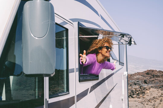 Cheerful adult woman open arms outstretching outside the window of her modern van camper admiring and enjoying travel destination and alternative home van life. Happy people smile in summer vacation