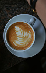 Cafe latte with leave foam print on a marble counter with cropped woman’s hand