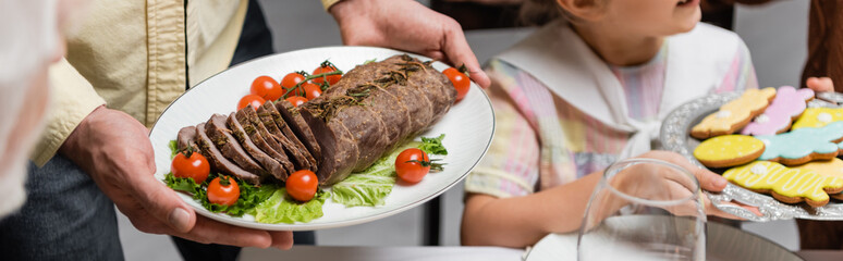 partial view of man holding plated with meat, cherry tomatoes and lettuce near daughter with cookies, banner.