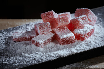 Traditional turkish delight lokum sugar coated soft candy. Red marmalade jelly candy's covered with powder sugar on a wooden cutting board on black background.