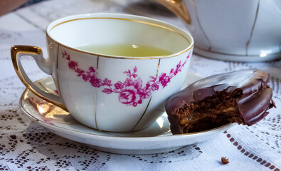 Porcelain tea cup with mint tea, chocolate and dulce de leche alfajor bitten on a napkin, on a table with white lace tablecloth.