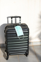 Black cabin-sized suitcase with a medical face mask with palm tree shadow on sunny day