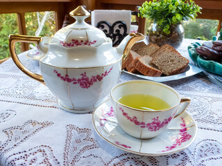 Porcelain teapot, tea cup with mint tea, slices of cake, alfajores, napkin and potted plant on a table with white lace tablecloth