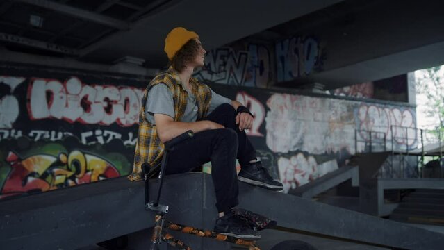 Calm teenager relaxing on ramp at skatepark. Handsome man having break.