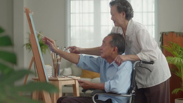 An elderly man in a wheelchair with his Asian wife is happy together painting with watercolors on vacation at home, everyone is happy and laughing. Concentrate on drawing and appreciating your work