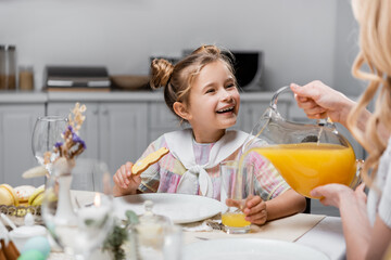 joyful girl holding traditional easter cookie near mom pouring fresh orange juice.