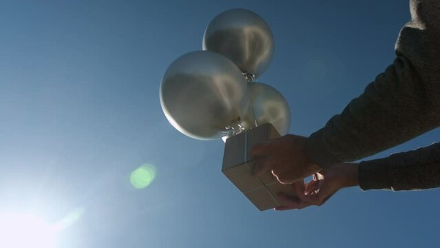 Man caught gift box attached to helium balloons at sunlight. Hands of person holding case against blue sky lit by sun rays. Low angle shot super slow motion