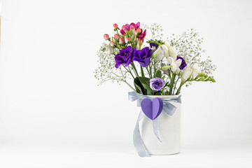 Different colors beautiful flowers bouquet in vintage white vase with ribbon and paper heart on white background. Romantic holiday background with copy space.