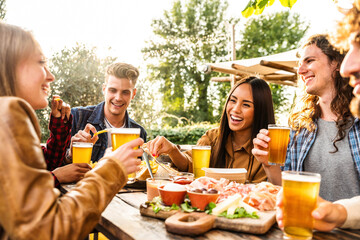 Group of multi ethnic friends having backyard dinner party together -  Diverse young people sitting at bar table toasting beer glasses in brewery pub garden - Happy hour, lunch break and youth concept