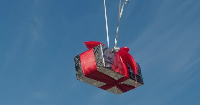 Silver present box attached to balloons floats in air. Shining case with decorative red ribbon reflects bright sunlight against blue sky closeup