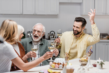 excited man gesturing with raised hand near family toasting with wine glasses.