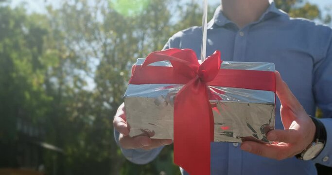 Man holds silver gift box with red ribbon attached to balloons on sunny day. Person shows present to camera standing against trees closeup slow motion