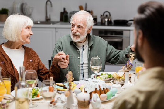 Happy Senior Couple Holding Hands During Easter Dinner With Blurred Son.