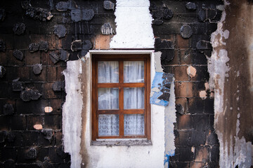 Old vintage wooden window with white curtains on burned out facade. Soot and char around window. Fire damaged bricks on house.