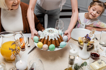 woman holding plate with painted eggs and easter cake near happy daughter and senior mother.