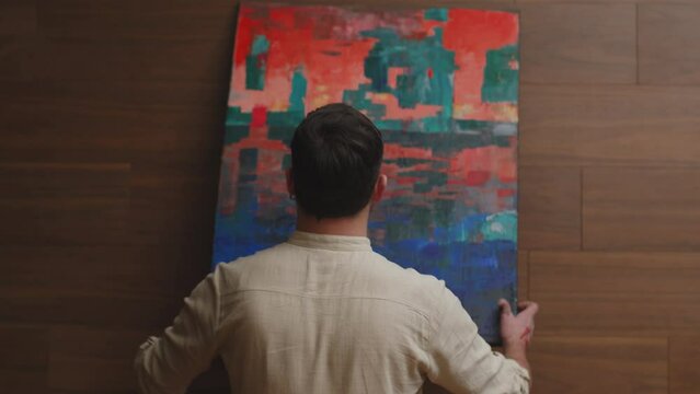 Contemporary art. Stylish guy artist hang colourful abstract artwork on wall, rear view, handheld video. ProRes codec