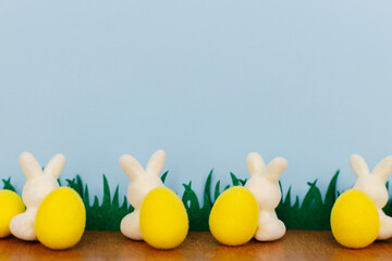 Happy Easter! Colorful Easter bunnies and eggs in grass on blue background with space for text. White and yellow artificial decor. Easter hunt concept