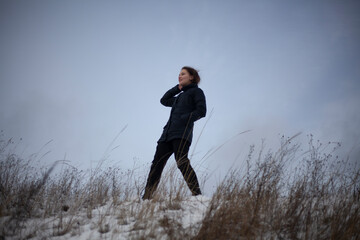 A girl stands in a field in winter. A young lady in black clothes is walking on the street. The model is removed in the open air. Teenage child. Overcast weather.