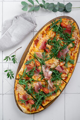 A long pizza with prosciutto, tomatoes, arugula and garlic on a tiled white tile. Italian dish...