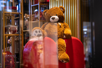 Large Fluffy teddy bear toy sits on big red heart gift box behind glass in the shop. Valentine's...
