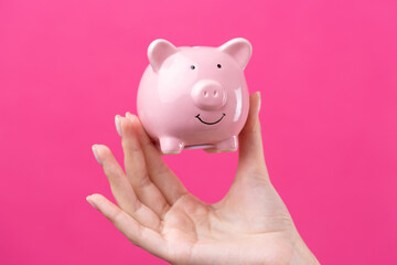 A cute girl in a pink jacket is holding a 3d piggy bank in her hands. Beautiful close-up portrait in studio on pink.