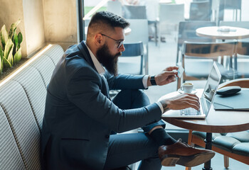 Serious Businessman Making a Credit Card Purchase while Sitting in a Cafe. 
Handsome businessman with beard and eyeglasses using his laptop computer for money transfer or cryptocurrency investment.