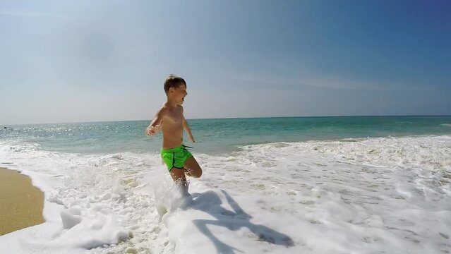 Slow motion little boy running along beach with waves and foam getting in the way and interrupting his run. Childhood, sea vacation concept.
