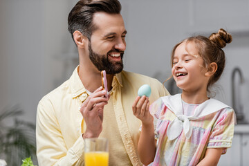 cheerful man and daughter holding cookie and painted egg during easter celebration.