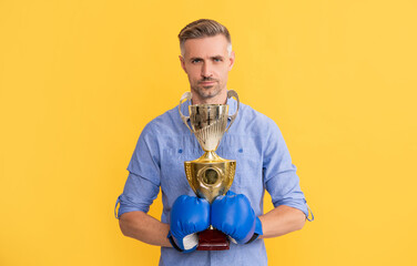 mature man in boxing gloves hold champion cup on yellow background, success