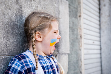 Portrait of a little girl who is screaming and with flag painted on her cheek in the yellow-blue colors of the Ukrainian flag. Concept of peace and protection of children