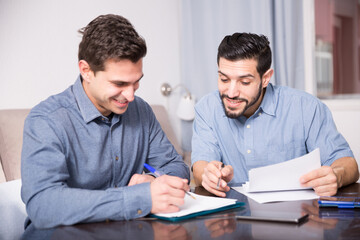 Portrait of two cheerful young men reading documents together at home table