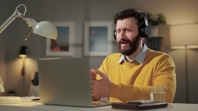 Man is embarrassed, shy emotion. Slightly confused bearded man with headphones in home office shyly and uncertainly looks into computer webcam
